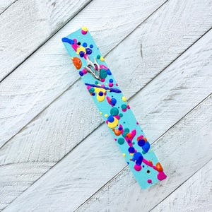 May include: A turquoise blue mezuzah with colorful paint splatters and a silver Star of David. The mezuzah is a decorative item for a Jewish home.