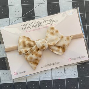 May include: A beige and brown bow with a matzah pattern on a nude elastic headband. The headband is packaged in a clear plastic bag with a white card that reads "Little Natalie Designs, llc handmade with love" and includes the website address and social media handles.