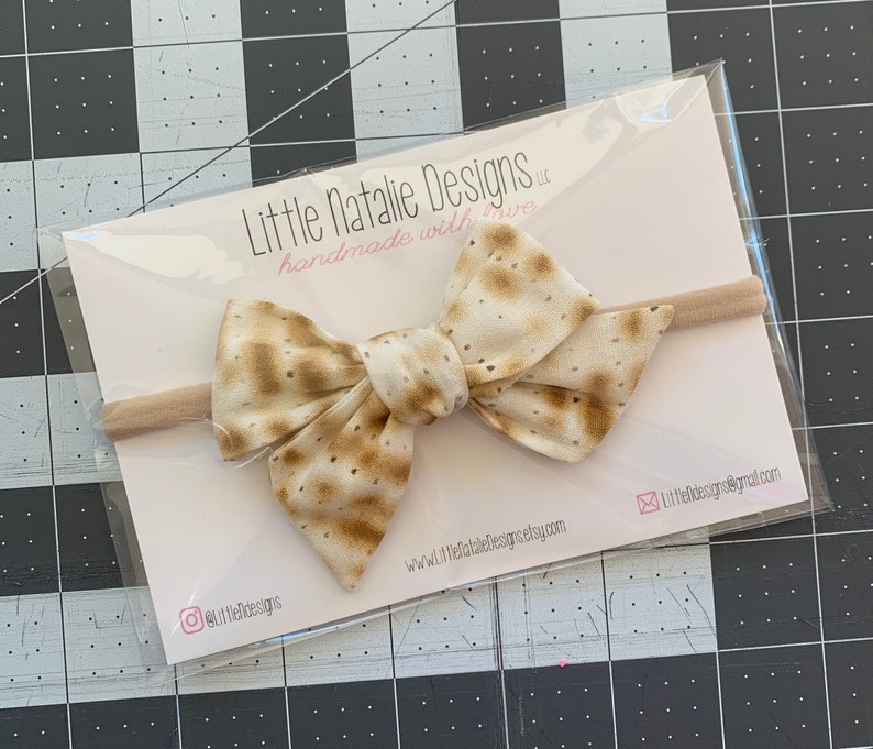 May include: A beige and brown bow with a matzah pattern on a nude elastic headband. The headband is packaged in a clear plastic bag with a white card that reads "Little Natalie Designs, llc handmade with love" and includes the website address and social media handles.
