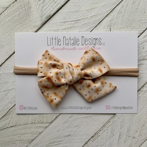 May include: A beige headband with a bow made from fabric printed with a matzah pattern. The headband is on a white background with the text "Little Natalie Designs LLC handmade with love" and contact information.