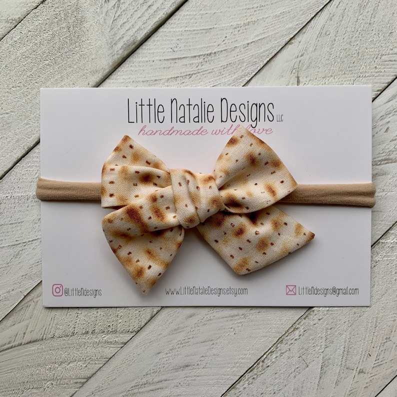 May include: A beige headband with a bow made from fabric printed with a matzah pattern. The headband is on a white background with the text "Little Natalie Designs LLC handmade with love" and contact information.