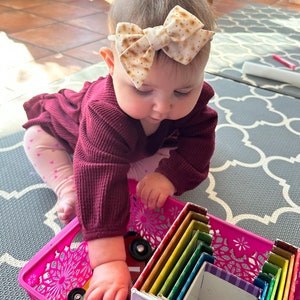 May include: A baby wearing a burgundy sweater and a headband with a brown and white bow is playing with a set of colorful nesting boxes. The boxes are stacked inside a pink plastic basket.