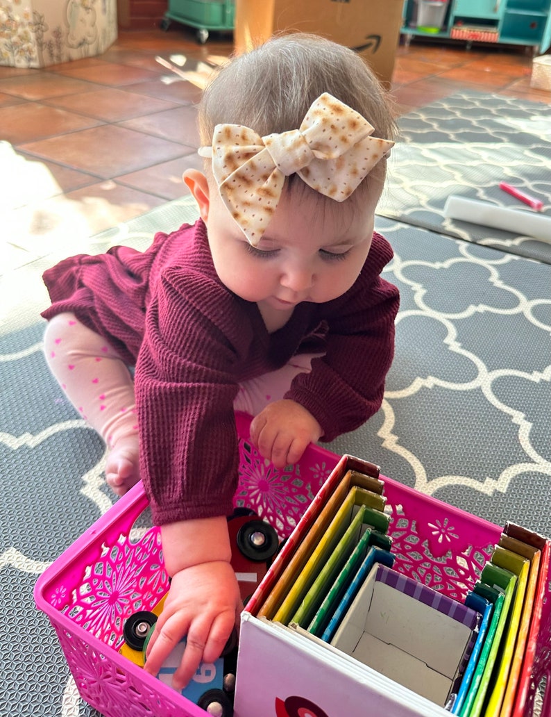 May include: A baby wearing a burgundy sweater and a headband with a brown and white bow is playing with a set of colorful nesting boxes. The boxes are stacked inside a pink plastic basket.