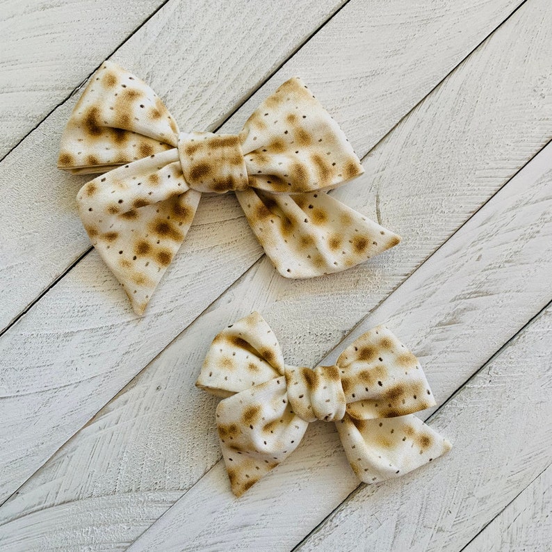 May include: Two fabric hair bows with a matzah pattern. The larger bow is brown and white with a more defined pattern. The smaller bow is a lighter brown and white with a less defined pattern.