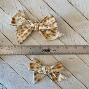 May include: Two beige and brown bow hair clips with a matzah pattern. The larger bow is about 4 inches wide and the smaller bow is about 2 inches wide.