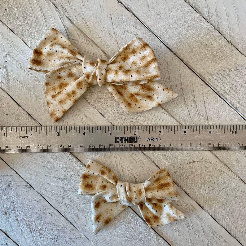 May include: Two beige and brown bow hair clips with a matzah pattern. The larger bow is about 4 inches wide and the smaller bow is about 2 inches wide.
