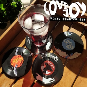 May include: Set of four black vinyl record coasters with colorful designs. The coasters feature a cat, a skeleton, a bird, and a cloud design. The text "Lovejoy" is featured on each coaster. The text "Design by Scivious" is printed on one of the coasters.