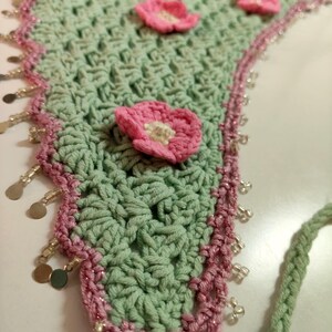 May include: A triangular crochet bandana in sage green featuring several small pink crocheted flowers with white centers. The edges are trimmed with a pink border, clear glass beads, and small metallic gold-toned circular charms.