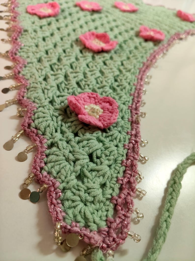 May include: A triangular crochet bandana in sage green featuring several small pink crocheted flowers with white centers. The edges are trimmed with a pink border, clear glass beads, and small metallic gold-toned circular charms.