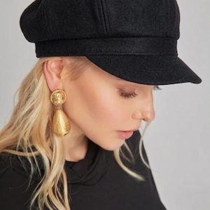 May include: A black wool newsboy cap with a short brim. The cap is worn with gold-coloured teardrop earrings. The model is wearing a black top.