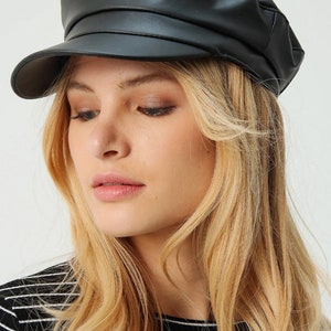 May include: A black faux leather baker boy hat with a rounded crown and a short, stiff brim. The hat is worn by a person with blonde hair and a black and white striped shirt.