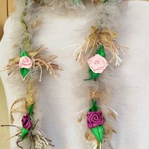 May include: A white and brown garland with pink and purple fabric flowers. The garland is made of tulle, burlap, and ribbon.