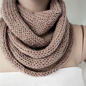以下が含まれることがあります： ニュートラルなダスティローズ色のニットインフィニティスカーフ。厚手のテクスチャード加工の糸でできており、ニットの模様が見えます。マネキンの首に巻かれており、暖かく居心地の良いデザインです。