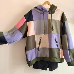 May include: A long-sleeve hooded sweatshirt featuring a patchwork design of rectangular panels in olive green, lavender, light pink, navy blue, and beige. The hoodie includes a front kangaroo pocket with two zippered openings and olive green drawstrings.