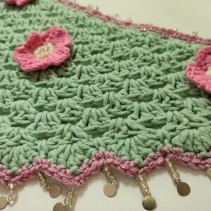 May include: A light green crocheted fabric featuring a repeating shell stitch pattern, adorned with three-dimensional pink flowers with white centers. The scalloped edge is trimmed in pink yarn with dangling clear beads and small metallic discs.