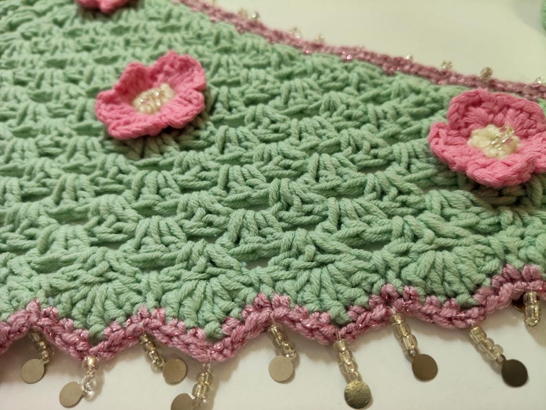May include: A light green crocheted fabric featuring a repeating shell stitch pattern, adorned with three-dimensional pink flowers with white centers. The scalloped edge is trimmed in pink yarn with dangling clear beads and small metallic discs.