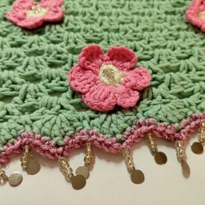 May include: A light green crocheted fabric featuring a repeating shell stitch pattern, adorned with three-dimensional pink flowers with cream centers. The scalloped edge is trimmed with a pink border and dangling clear beads and gold discs.