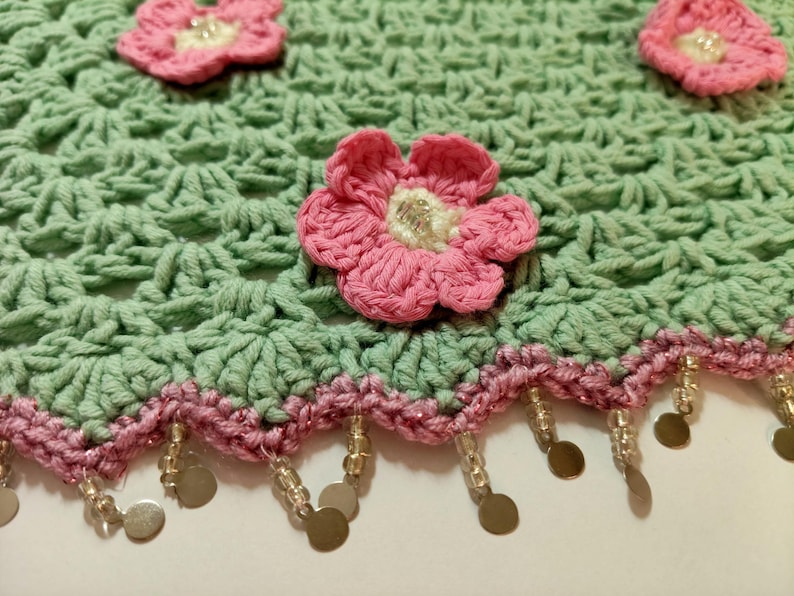 May include: A light green crocheted fabric featuring a repeating shell stitch pattern, adorned with three-dimensional pink flowers with cream centers. The scalloped edge is trimmed with a pink border and dangling clear beads and gold discs.