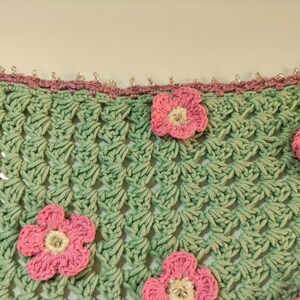 May include: A light green crochet fabric featuring a repeating shell stitch pattern. The piece is adorned with several five-petaled pink flowers with white centers and small clear beads. A mauve crochet border with clear beads lines the top edge.
