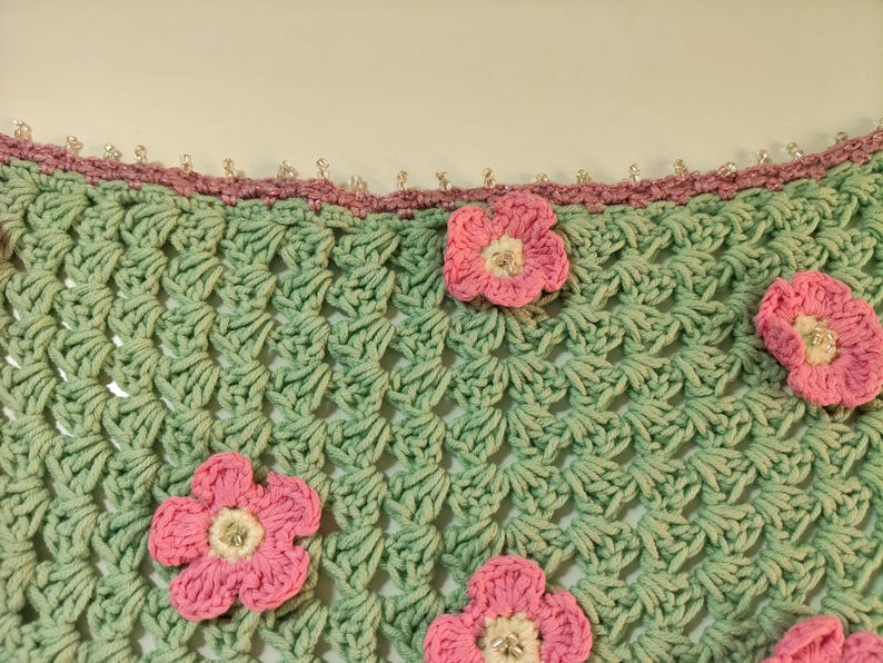 May include: A light green crochet fabric featuring a repeating shell stitch pattern. The piece is adorned with several five-petaled pink flowers with white centers and small clear beads. A mauve crochet border with clear beads lines the top edge.