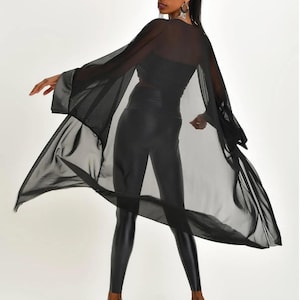 May include: A black sheer cape with wide sleeves and a satin trim. The cape is paired with black leggings and silver high heels. The model is wearing a black top.