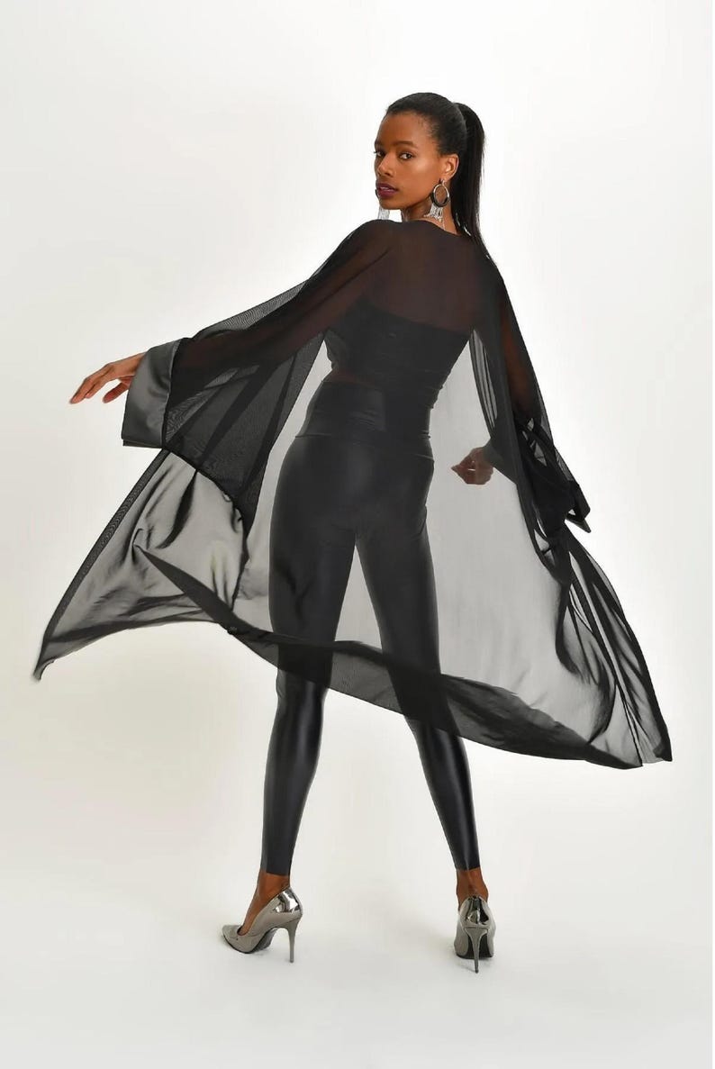 May include: A black sheer cape with wide sleeves and a satin trim. The cape is paired with black leggings and silver high heels. The model is wearing a black top.