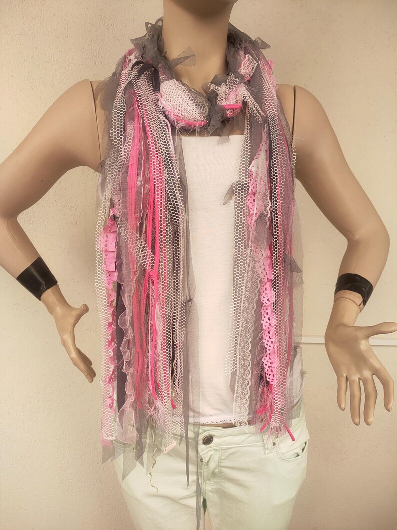 Op de afbeelding: Een grijze, roze en witte sjaal gemaakt van tule en kant. De sjaal heeft een rafelige rand en is om de nek van een mannequin geknoopt.