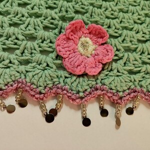 May include: A light green crocheted fabric featuring a shell stitch pattern, adorned with a pink crocheted flower with a white center. The bottom edge is finished with a pink crocheted border and dangling clear beads and small metallic discs.