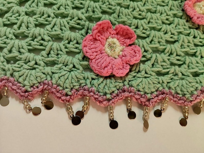 May include: A light green crocheted fabric featuring a shell stitch pattern, adorned with a pink crocheted flower with a white center. The bottom edge is finished with a pink crocheted border and dangling clear beads and small metallic discs.