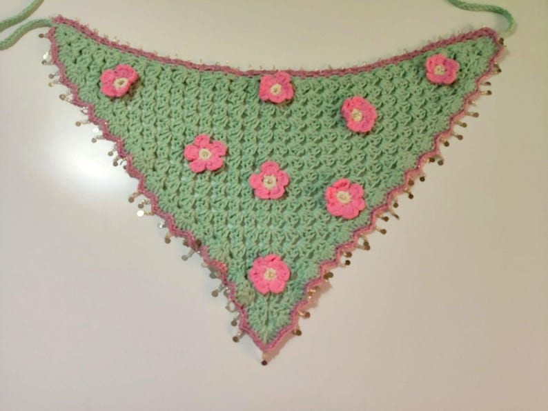 May include: A triangular crochet bandana in light mint green featuring a pink crocheted border and six small pink floral appliqu&eacute;s scattered across the surface. The edges are adorned with small, dangling metallic silver sequins.