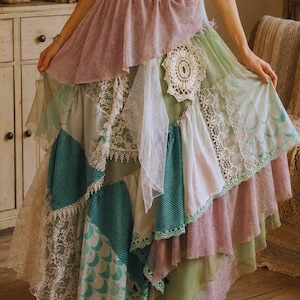 May include: A long, layered skirt featuring a patchwork design of various fabrics including sage green, teal, and lavender. The skirt incorporates white lace panels, a circular crochet doily accent, and a teal fabric with a crescent moon pattern.