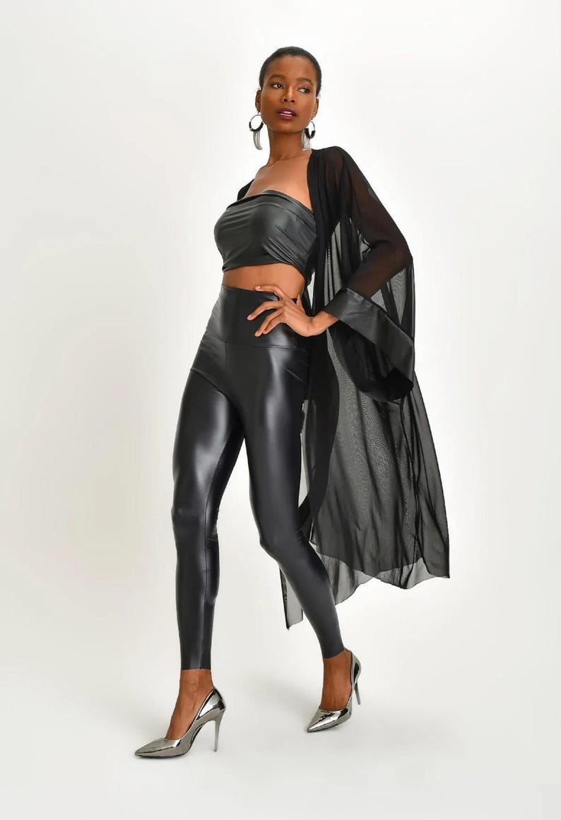 May include: A woman's ensemble featuring a black faux leather tube top, high-waisted leggings, and a sheer black kimono-style cover-up. The outfit is completed with silver high-heeled shoes. The overall look is sleek and modern.