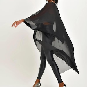 May include: A woman wearing a flowing black sheer cape with silver trim, black leggings, and silver high heels. The cape drapes over the shoulders and arms, creating a dynamic silhouette. The outfit is set against a white background.