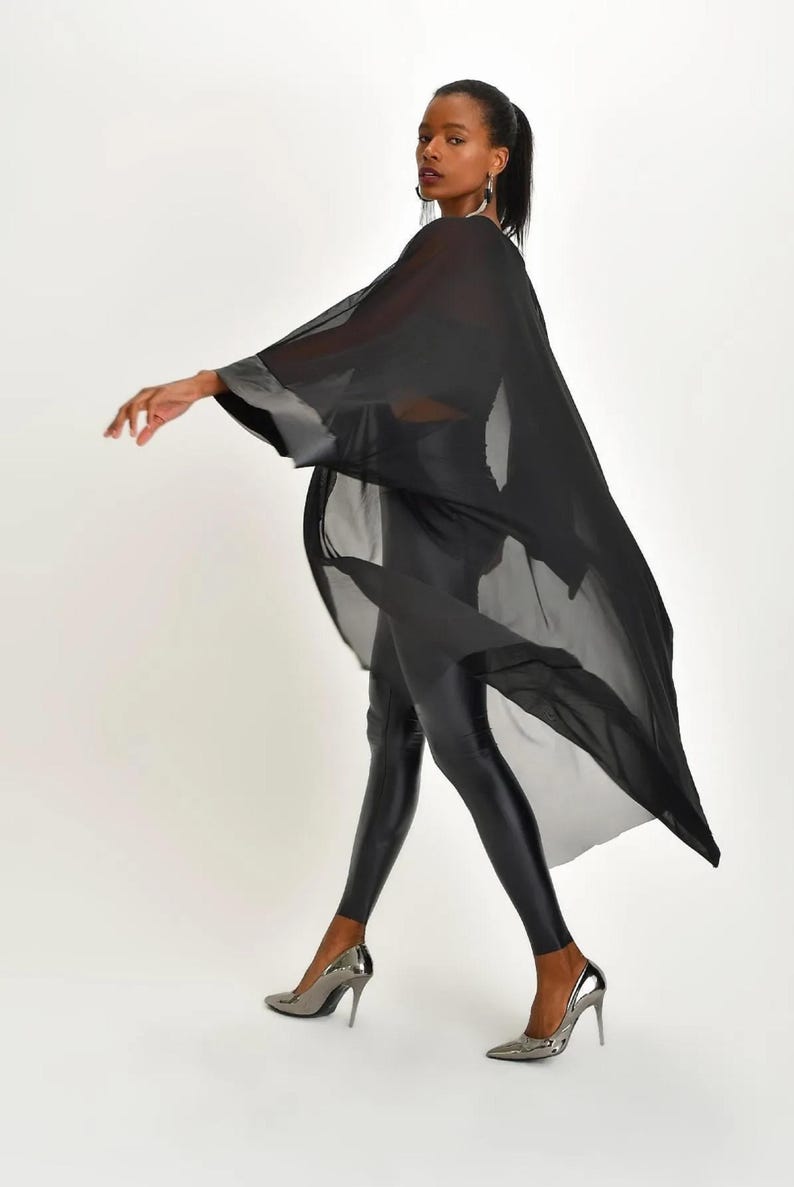 May include: A woman wearing a flowing black sheer cape with silver trim, black leggings, and silver high heels. The cape drapes over the shoulders and arms, creating a dynamic silhouette. The outfit is set against a white background.