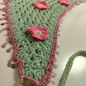 May include: A triangular crochet bandana in light sage green featuring several small pink flowers with white centers attached to the surface. The edges are trimmed with a pink border, clear glass beads, and small metallic gold circular charms.