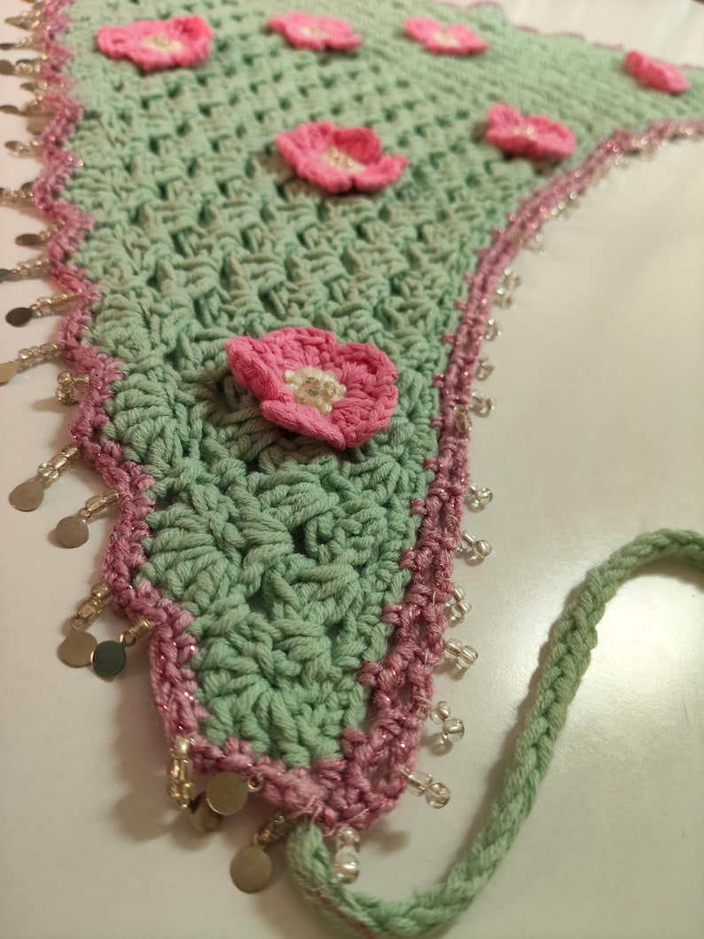 May include: A triangular crochet bandana in light sage green featuring several small pink flowers with white centers attached to the surface. The edges are trimmed with a pink border, clear glass beads, and small metallic gold circular charms.