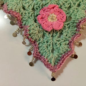 May include: A triangular crochet shawl featuring a light mint green openwork pattern with a pink crocheted flower at the center. The edges are trimmed with a pink border and decorated with dangling clear beads and small metallic gold circular discs.