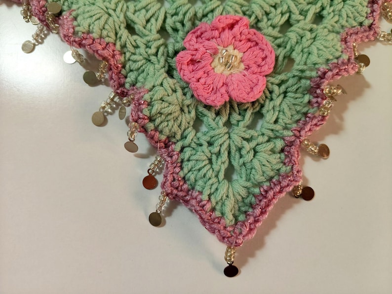 May include: A triangular crochet shawl featuring a light mint green openwork pattern with a pink crocheted flower at the center. The edges are trimmed with a pink border and decorated with dangling clear beads and small metallic gold circular discs.