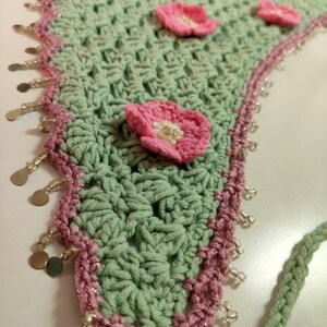 May include: A triangular crochet bandana in sage green featuring several small pink floral appliqu&eacute;s with white centers. The edges are trimmed in a darker pink yarn with a decorative border of dangling silver-toned metal discs and clear beads.
