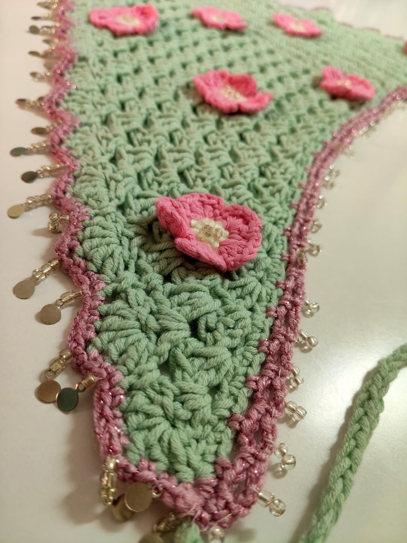 May include: A triangular crochet bandana in sage green featuring several small pink floral appliqu&eacute;s with white centers. The edges are trimmed in a darker pink yarn with a decorative border of dangling silver-toned metal discs and clear beads.