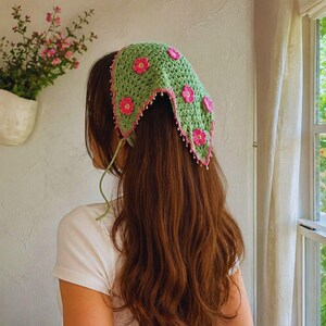 May include: A triangular crochet headscarf in light green featuring pink floral appliqu&eacute;s and a pink scalloped border with small white beads along the edges. The headscarf is tied with thin green strings at the back of the head.