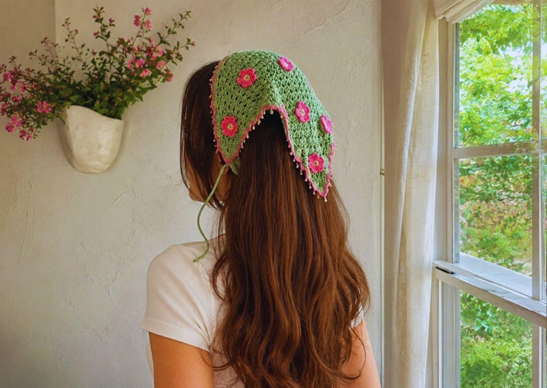 May include: A triangular crochet headscarf in light green featuring pink floral appliqu&eacute;s and a pink scalloped border with small white beads along the edges. The headscarf is tied with thin green strings at the back of the head.