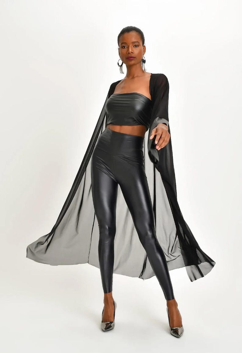 May include: A black ensemble featuring a strapless crop top, high-waisted leggings, and a sheer, flowing cape. The outfit is completed with pointed-toe heels. The clothing has a shiny, leather-like appearance.