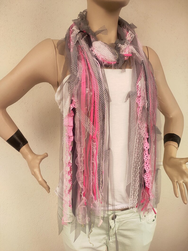 Op de afbeelding: Een grijze, roze en witte sjaal gemaakt van gelaagd tule, kant en lint. De sjaal is gescheurd en heeft een verweerde look.