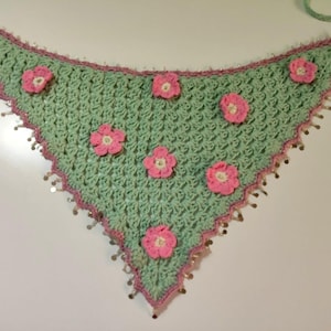 May include: A triangular crochet bandana in light mint green featuring a textured stitch pattern. The edges are trimmed in a darker pink yarn with small metallic silver sequins attached. Six small pink crocheted flowers are sewn onto the surface.