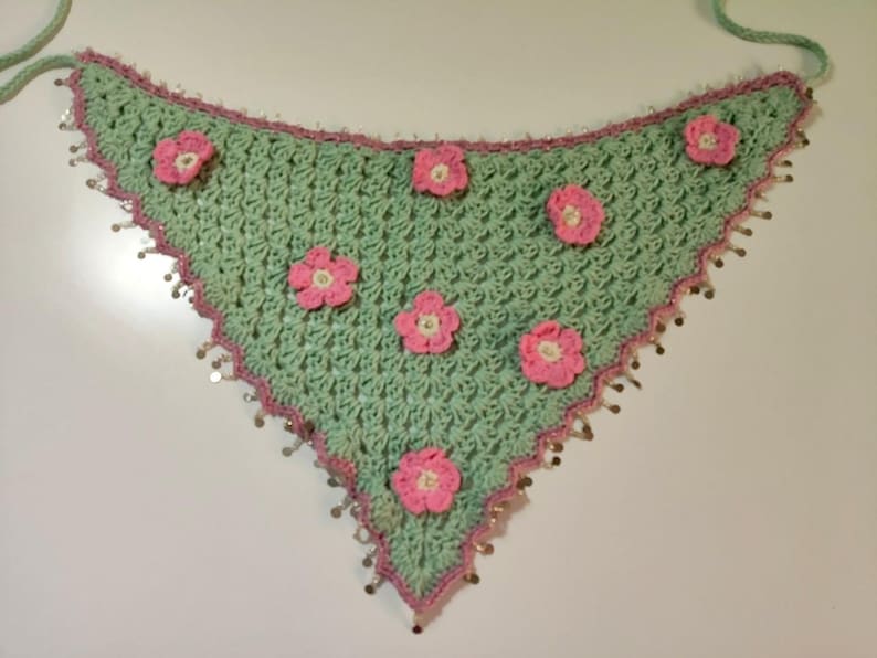 May include: A triangular crochet bandana in light mint green featuring a textured stitch pattern. The edges are trimmed in a darker pink yarn with small metallic silver sequins attached. Six small pink crocheted flowers are sewn onto the surface.