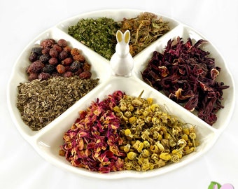 May include: A white ceramic plate divided into six sections, each filled with a different type of dried herb. The herbs include rose petals, chamomile flowers, hibiscus flowers, dried mint leaves, and rose hips.