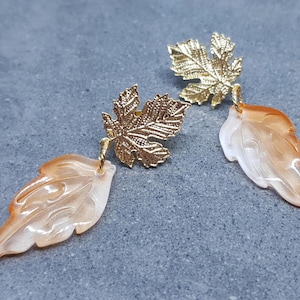 Op de afbeelding: Een paar gouden bladeren oorbellen met een perzikkleurige schelp blad hanger. Het schelp blad heeft een gedetailleerd gesneden ontwerp.