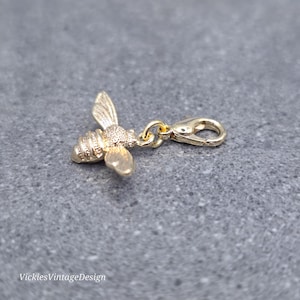 Puede incluir: Un dije de abeja de color dorado con un cierre de mosquetón. La abeja tiene alas y segmentos corporales detallados. El dije probablemente está hecho de metal y está diseñado para sujetarse a una pulsera o collar. El dije está sobre una superficie gris.
