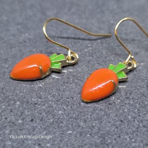 Könnte beinhalten: Ein Paar goldfarbene Karottenohrringe. Jeder Ohrring zeigt eine leuchtend orangefarbene Karotte mit grünen Blättern. Die Ohrringe haben einen goldfarbenen Haken für durchbohrte Ohren.
